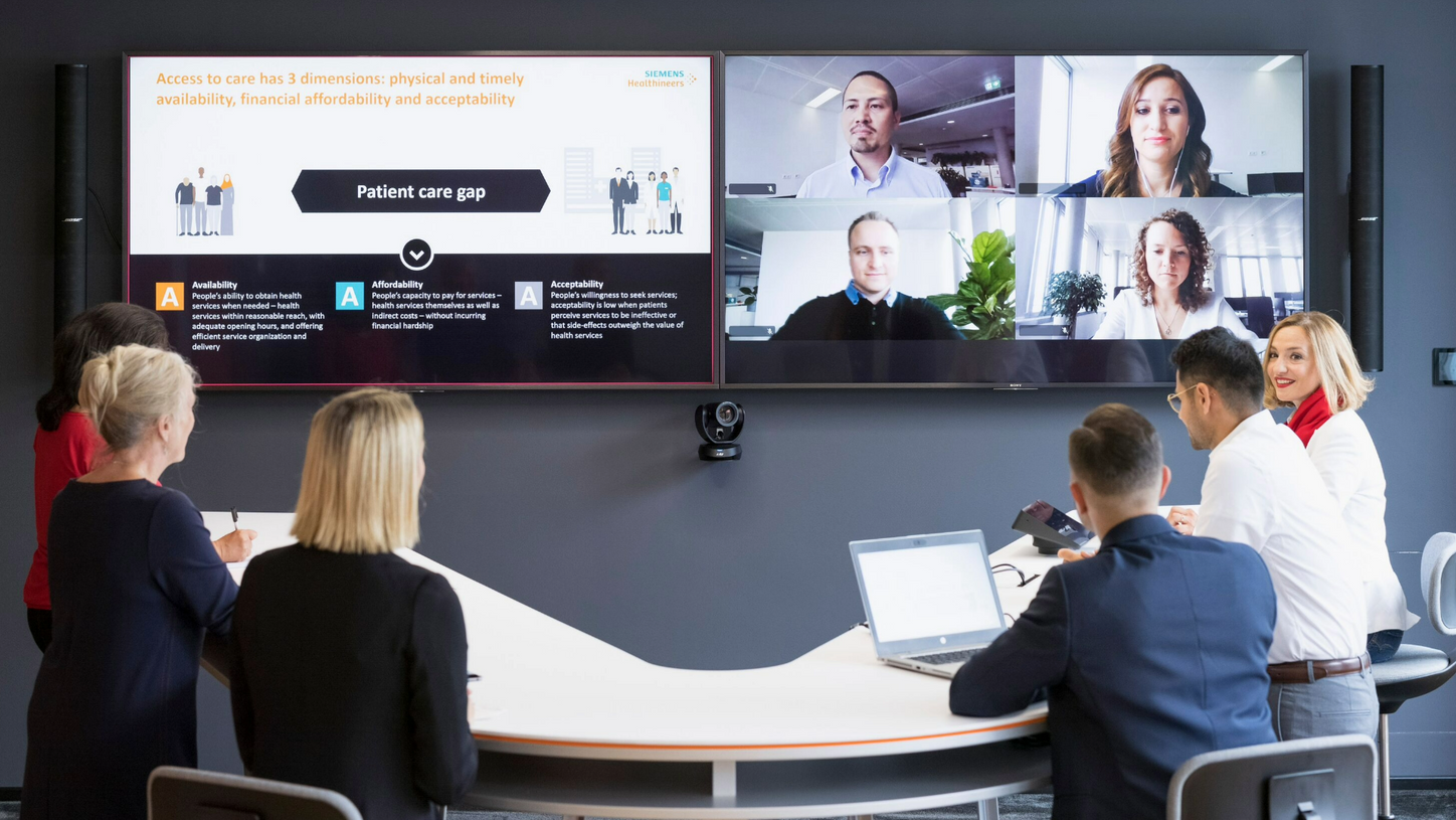 Kolleg*innen in Meetingraum nehmen an einem hybriden Meeting mit Mitarbeitenden im Videocall teil.