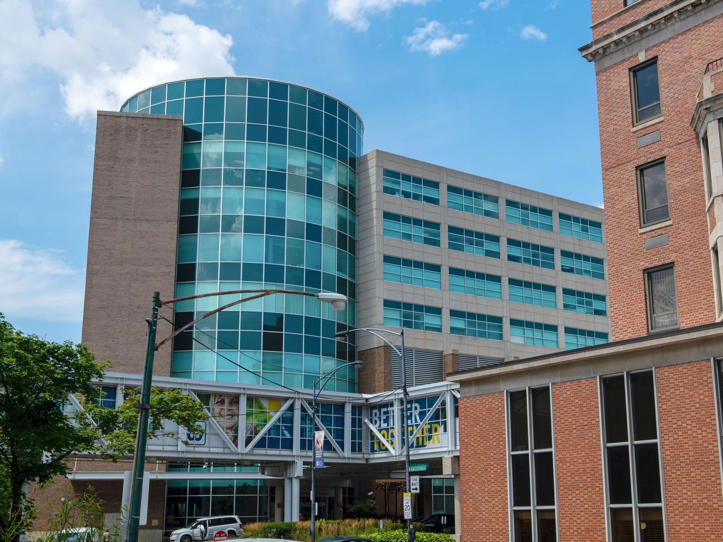 Exterior of a hospital building featuring a cylindrical glass tower and an enclosed pedestrian walkway.
