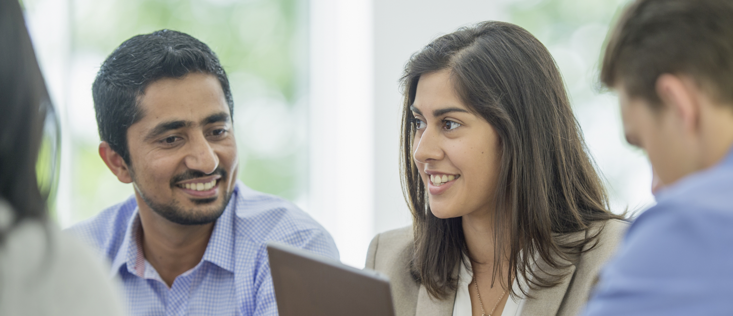 A multi-ethnic group of young business men and women in semi-casual office clothes are sharing ideas and holding a digital tablet in an indoor, sunlit office