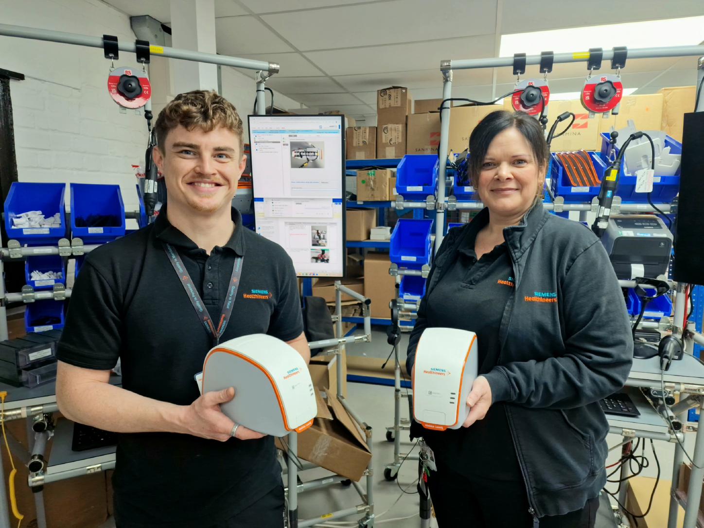 Two members of the Siemens Healthineers Sudbury manufacturing team holding the Atellica LumIQ Point-of-Care Analyser, showcasing the advanced, compact device designed for modern urinalysis testing. 