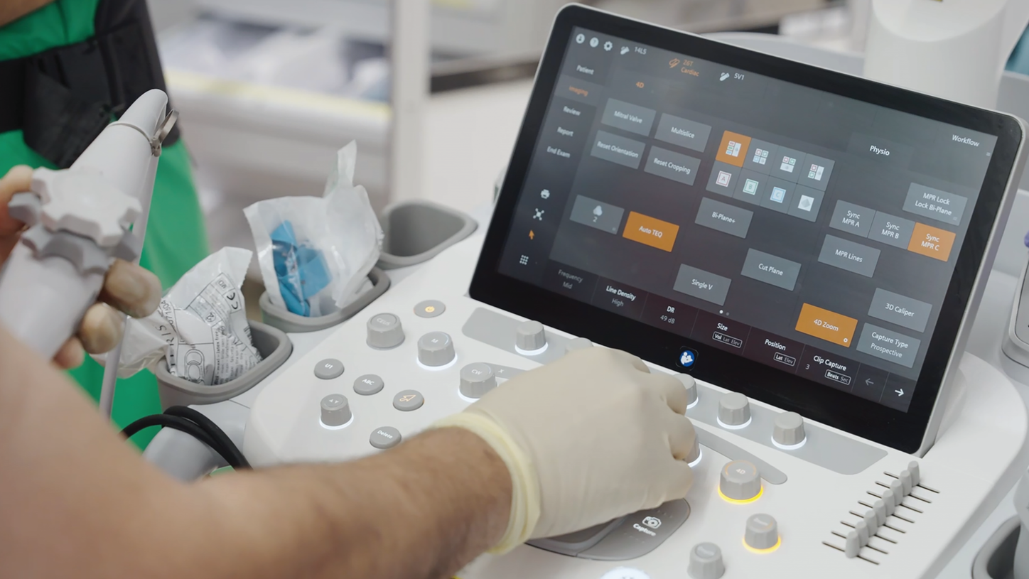 close-up of a gloved hand interacting with a dark-themed touchscreen interface mounted on ACUSON Origin; the screen shows a grid of control icons and status indicators.