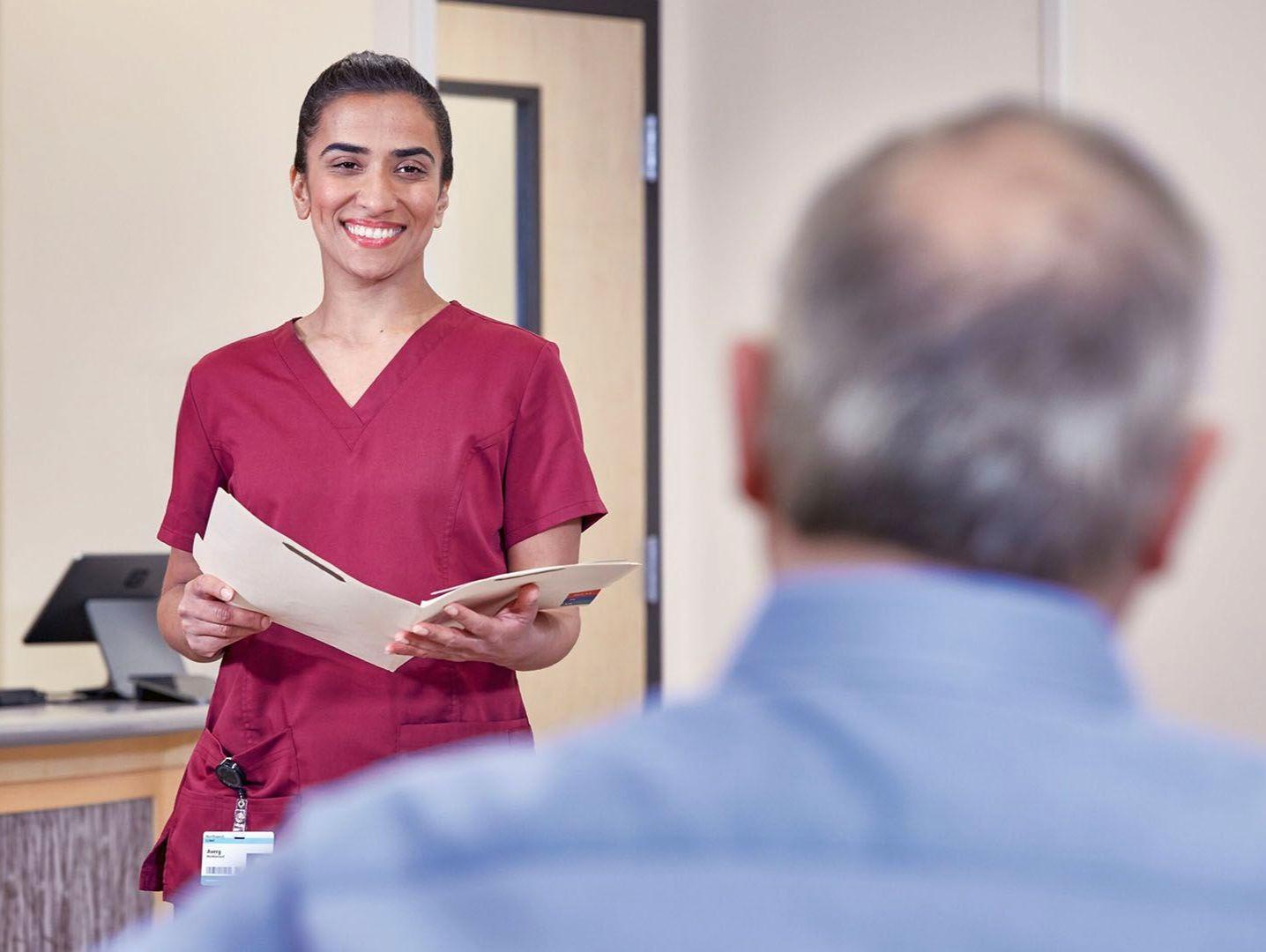 A nurse wearing pink scrubs welcomes an older male patient in the waiting room holding his chart