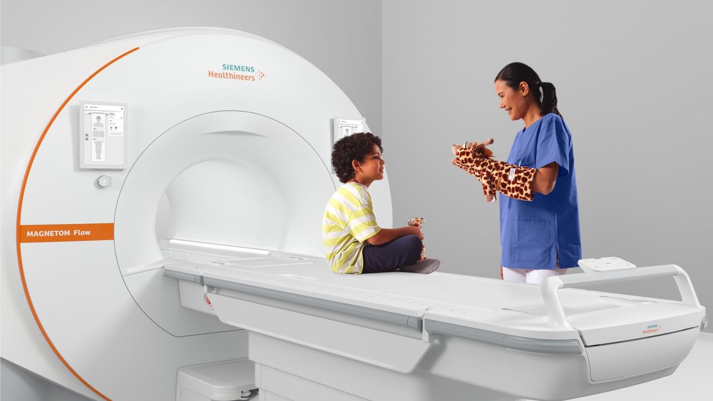 A child sits on an MRI scanner table holding a stuffed giraffe while a healthcare professional in blue scrubs uses a giraffe hand puppet to engage and comfort them