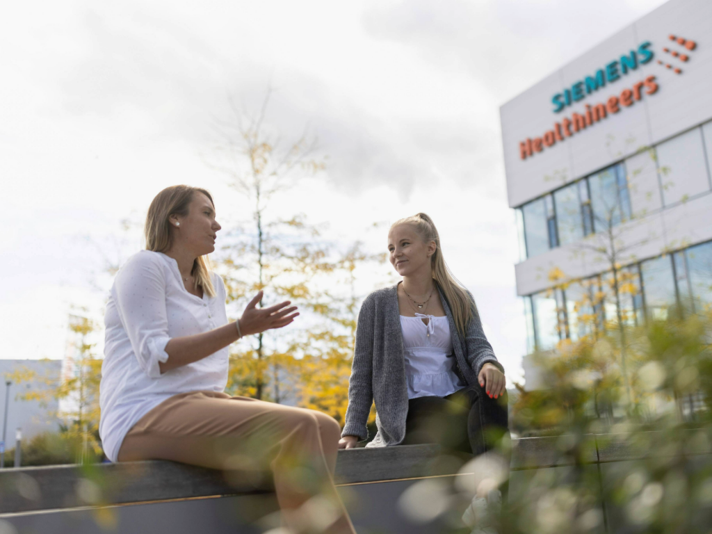 Zwei weibliche Mitarbeitende tauschen sich vor dem Siemens Healthineers Bürogebäude aus.