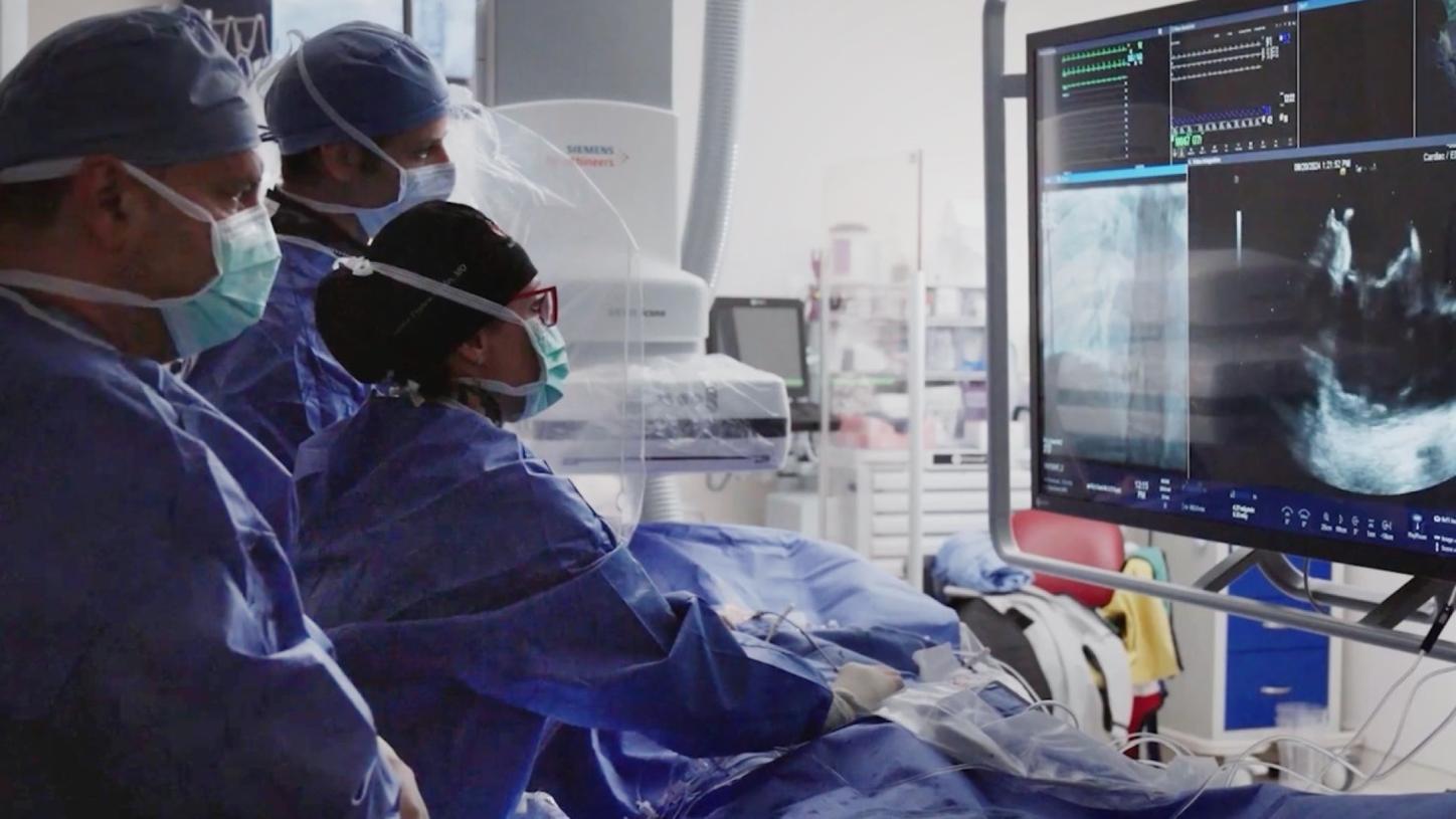 Three medical professionals in surgical attire are performing a procedure while observing real-time imaging on a monitor displaying ultrasound and vital signs.