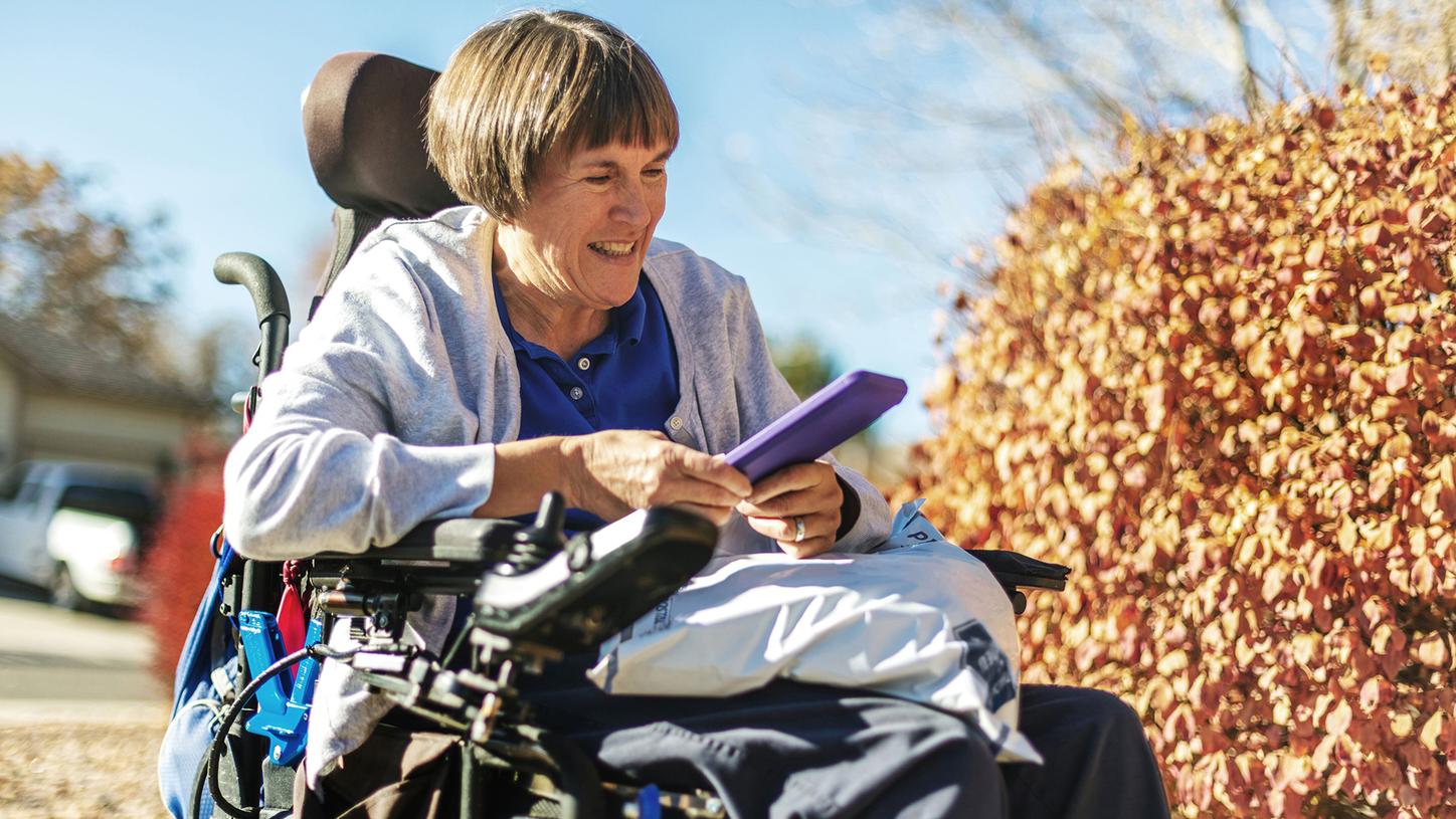 Woman using a powered wheelchair outdoors while looking at a tablet device, with autumn foliage in the background.