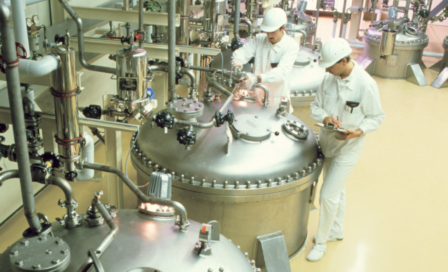  Technicians monitor and document large stainless-steel bioreactors connected by piping in a controlled laboratory environment for cell culture research.