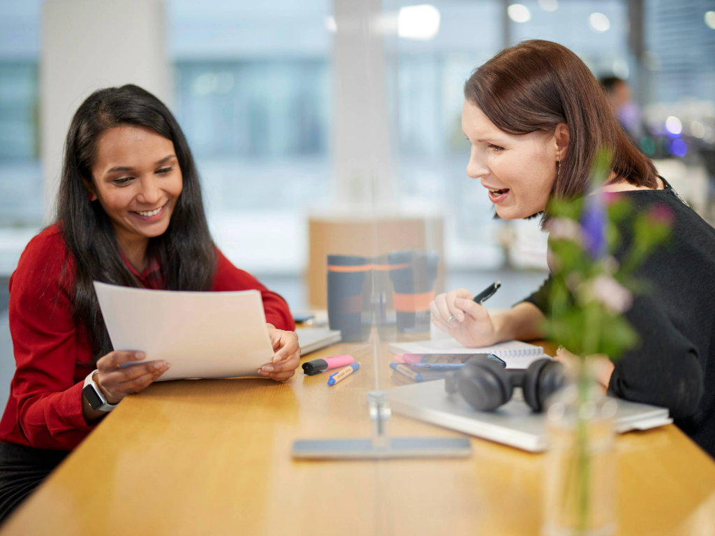 Zwei weibliche Kolleginnen an einem Stehtisch sprechen über ein Dokument.