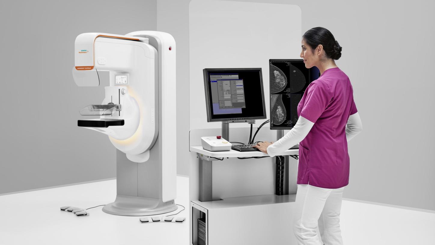 Technician operating a mammography system while reviewing breast images on dual monitors in a clinical setting.